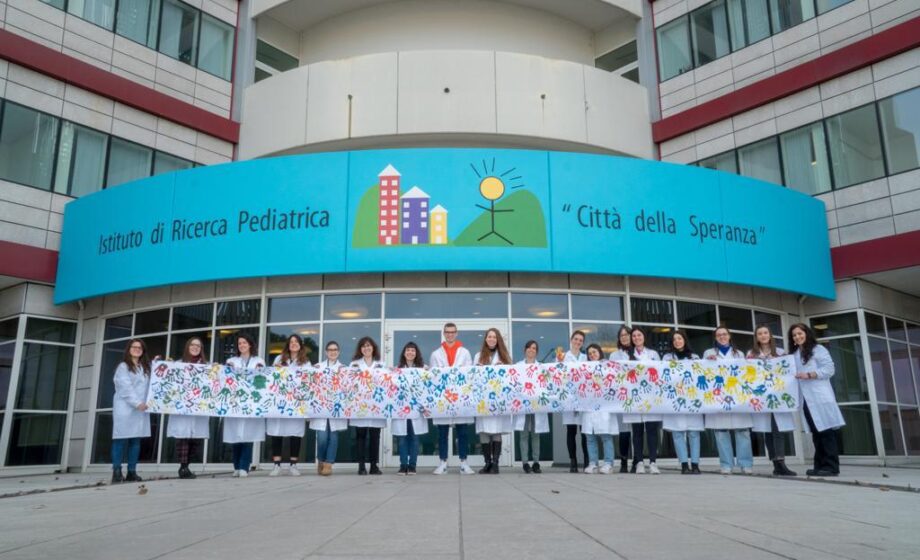 Foto dell'ingresso della Città Della Speranza con decine di ricercatori che tengono in mano un lenzuolo bianco in cui sono impresse le impronte delle mani, di diversi colori, di molti bambini