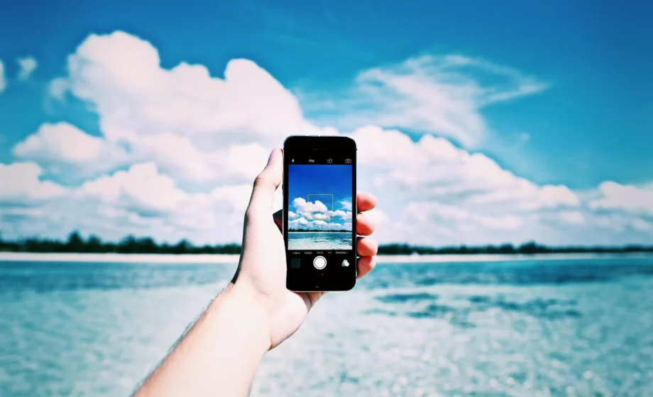 Una mano tiene uno smartphone che fotografa un cielo azzurro con grandi nuvole bianche sopra il mare, evocando una sensazione di calma, libertà e vero riposo estivo